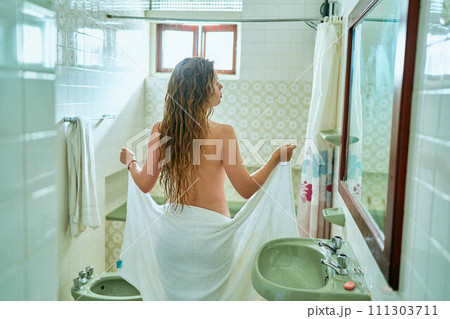Rear view of a young woman drying off with a towel in a vintage bathroom setting. Female post-bath routine, personal care in domestic interior with tiled walls, mirror, basin. 111303711