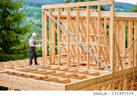 Carpenter building wooden skeleton house. Man measures distances with tape measure while wearing work attire and safety helmet. The principles of contemporary, eco-friendly building practices. Carpenter building wooden skeleton house. Man measures distances with tape measure while wearing work attire and safety helmet. The principles of contemporary, eco-friendly building practices. 111307324