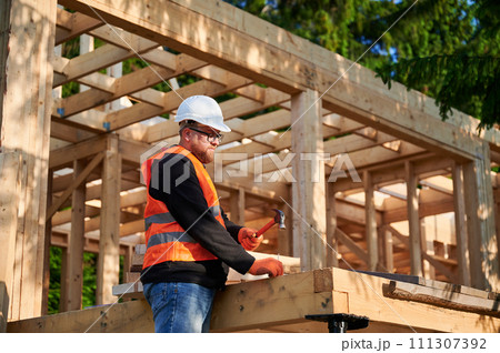 Carpenter constructing wooden frame house near forest. Bearded man with glasses hammering nails into structure, wearing protective helmet, construction vest. Concept of modern ecological construction. 111307392