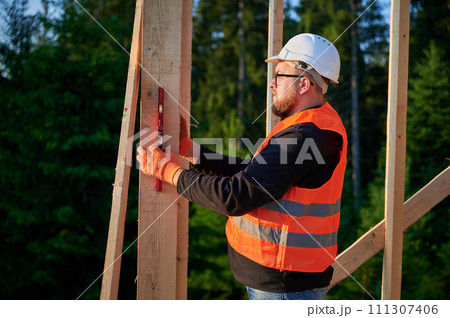 Carpenter constructing wooden frame house near the forest. Bearded man, wearing spectacles, inspects the walls for levelness using spirit level. He is attired in protective helmet, and orange vest. Carpenter constructing wooden frame house near the forest. Bearded man, wearing spectacles, inspects the walls for levelness using spirit level. He is attired in protective helmet, and orange vest. 111307406