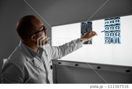 Side view of male doctor studying MRI results. Male specialist working in hospital, carrying out description of the radiographs of patient, pointing by pen. Specialist wearing glasses and white robe. 111307516