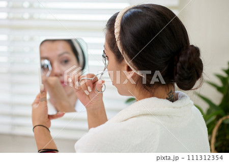 Brunette girl in white bathrobe curling eyelashes and looking in mirror in her hand while taking care of her beauty in bathroom 111312354