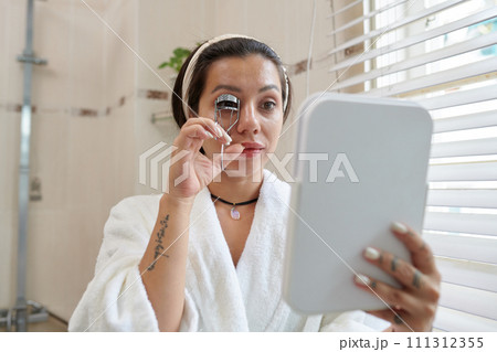 Young woman in soft white bathrobe looking in mirror in her hand and using eyelash curler while standing in bathroom 111312355