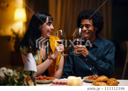 Happy young man and woman with glasses of red wine toasting for love and Valentine Day while sitting by served table at dinner 111312396