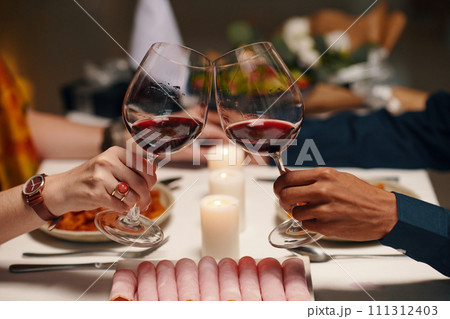 Hands of young unrecognizable sweethearts clinking with glasses of wine over table served with homemade food and fresh platter Hands of young unrecognizable sweethearts clinking with glasses of wine over table served with homemade food and fresh platter 111312403