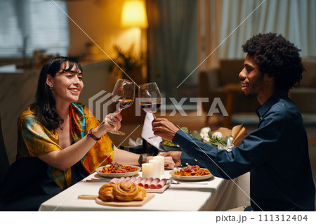 Happy young sweethearts toasting for love and clinking with glasses of red wine while sitting by served festive table and celebrating Valentine Day 111312404