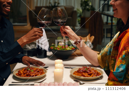 Cropped shot of happy young intercultural valentines clinking with glasses of red wine while sitting by served table with homemade food 111312406