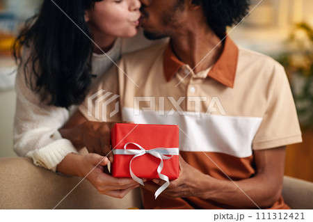 Hands of young affectionate husband and wife kissing each other while holding red giftbox with packed present for valentine day 111312421