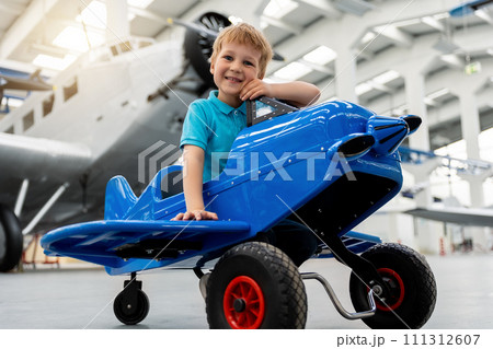 Portrait of cute little blond happy kid boy enjoy have fun play riding pedal toy plane model against vintage airplane background in museum hangar. Future child profession dream concept 111312607