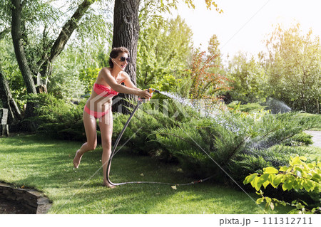 Young adult sexy slim sporty caucasian brunette woman enjoy having fun watering garden at home yard with hose pipe shower on hot summer day. Happy female person relax at backyard park outdoors 111312711