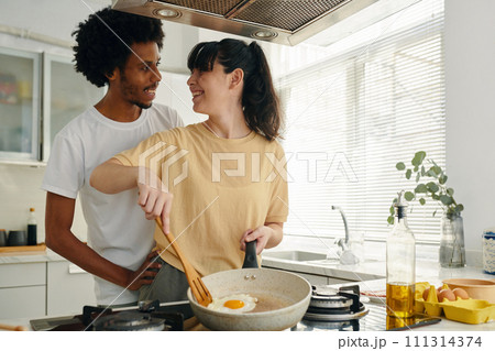 Pretty young woman with frying pan cooking scrambled eggs for breakfast while standing by electric stove and looking at her husband Pretty young woman with frying pan cooking scrambled eggs for breakfast while standing by electric stove and looking at her husband 111314374