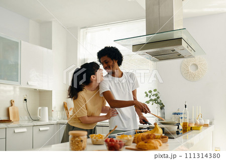 Young cheerful man looking at his wife while standing by electric stove in the kitchen and preparing something tasty for breakfast Young cheerful man looking at his wife while standing by electric stove in the kitchen and preparing something tasty for breakfast 111314380