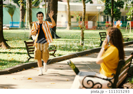 Young friendly guy with backpack waving hand to girl with laptop while walking along road in park on sunny summer day and Young friendly guy with backpack waving hand to girl with laptop while walking along road in park on sunny summer day and 111315293