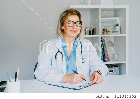 Confident mature middle aged woman doctor medical nurse, female physician practitioner in white coat with stethoscope sitting at working place at hospital office and making notes during appointment. Confident mature middle aged woman doctor medical nurse, female physician practitioner in white coat with stethoscope sitting at working place at hospital office and making notes during appointment. 111315302