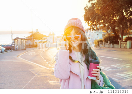 Young smiling hipster woman with color hair holding reusable coffee cup wearing pink coat, knitted hat and pink sunglasses on the street walk in sunset light, happy mood, seasonal fashion style trend 111315315