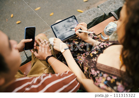 Hands of young man with smartphone and girl with stylus drawing sketch of nature on screen of tablet at leisure while sitting on bench 111315322