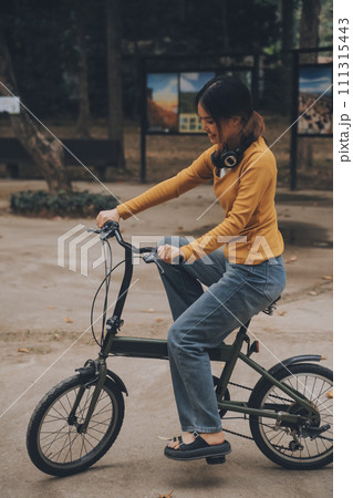 Happy young Asian woman while riding a bicycle in a city park. She smiled using the bicycle of transportation. Environmentally friendly concept. Happy young Asian woman while riding a bicycle in a city park. She smiled using the bicycle of transportation. Environmentally friendly concept. 111315443