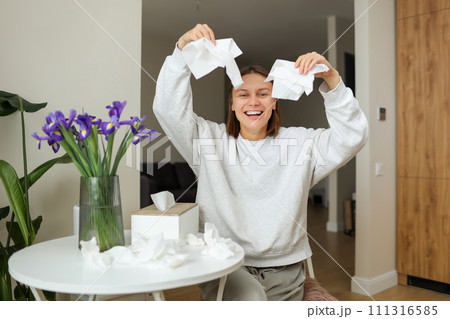 Seasonal allergy free. Happy young woman throws tissues up at home. Healthy life without of runny nose, itching or cough symptoms. Allergen bouquet of iris flowers at vase and used napkins on table. 111316585