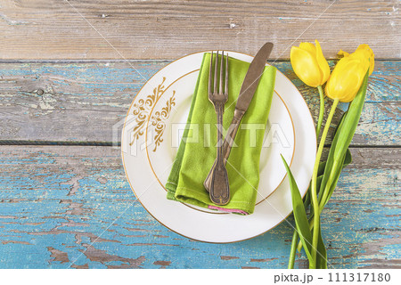 Happy easter holidays or valentines day concept. Top view for place setting with vintage fork and knife on a green napkin and two white plates, bouquet of yellow tulips on a old paint blue wooden Happy easter holidays or valentines day concept. Top view for place setting with vintage fork and knife on a green napkin and two white plates, bouquet of yellow tulips on a old paint blue wooden 111317180