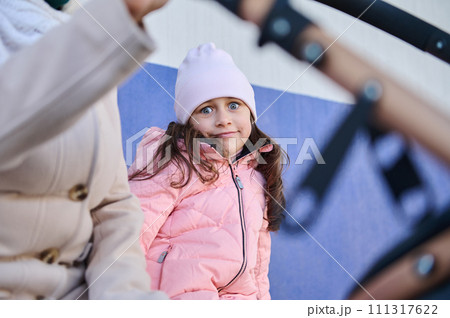 Little child girl in warm clothes, looking at camera, sitting on a banc on the railway station, waiting for urban train 111317622