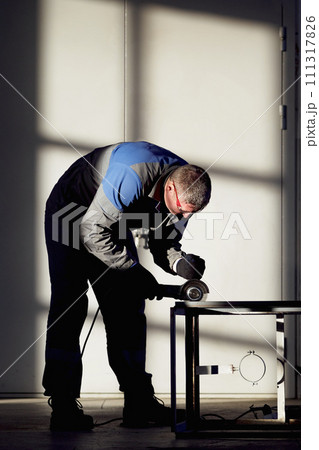 A skilled worker is shown focused on grinding a metal piece with a handheld power tool in a brightly A skilled worker is shown focused on grinding a metal piece with a handheld power tool in a brightly 111317826