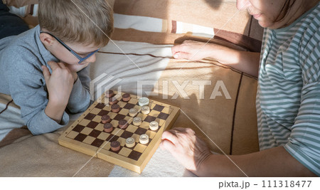 A caring middle-aged grandmother plays checkers with a child, a boy, lying on the bed. Happy family enjoying an interesting wooden board game at home. 111318477