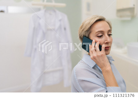 Medium shot of focused female doctor talking on smartphone in hospital clinic on background of white coat. Confident medical worker physician talks about problem to patient at workplace in office 111321376