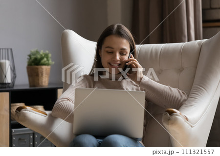 Young woman talks on smartphone seated on armchair with laptop 111321557