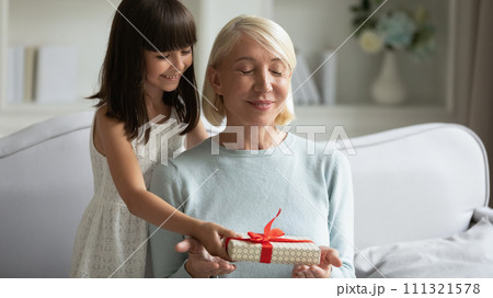 Little granddaughter preparing surprise for smiling mature grandmother, presenting gift 111321578