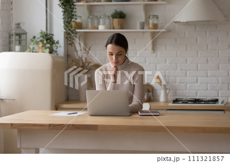 Serious woman sit at table in domestic kitchen with laptop 111321857