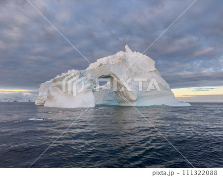 A huge high breakaway glacier drifts in the southern ocean off the coast of Antarctica at sunset, the Antarctic Peninsula, the Southern Arctic Circle, azure water, cloudy weather 111322088