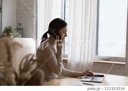 Woman talking on cellphone ordering goods use laptop at home 111323534