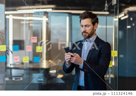 Serious businessman in suit using smartphone in modern office. Focused on finance, leadership, and corporate lifestyle. 111324146
