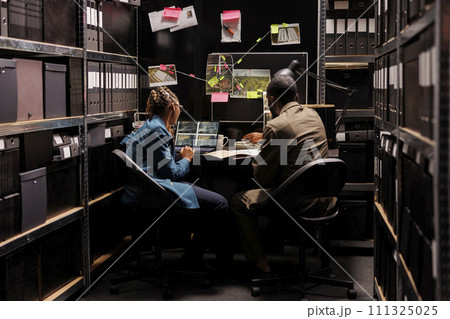 Private detectives looking at surveillance photos on laptop and analyzing report. African american man and woman investigators examining clues in dark agency office at night Private detectives looking at surveillance photos on laptop and analyzing report. African american man and woman investigators examining clues in dark agency office at night 111325025