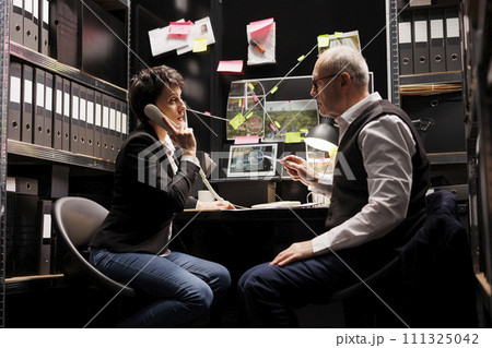 Private detective talking at landline phone with remote inspector, discussing missing person case. Senior police officer analyzing crime evidence, working late at night in arhive room 111325042