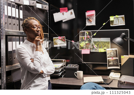 Pensive african american woman detective thinking about solving crime and rubbing chin. Thoughtful private investigator analyzing evidence while standing in agency office room Pensive african american woman detective thinking about solving crime and rubbing chin. Thoughtful private investigator analyzing evidence while standing in agency office room 111325043