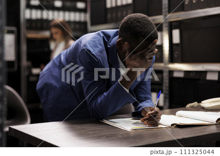 African american inspector thinking at ideas to solve criminal case, analyzing investigation report. Private detective searching for criminology evidence, working overtime in arhive room. 111325044