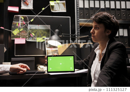 Private detectives working at criminal case in arhive room, discussing confidential information, analyzing scene evidence. Tablet computer with green screen chroma key mock up display standing on desk Private detectives working at criminal case in arhive room, discussing confidential information, analyzing scene evidence. Tablet computer with green screen chroma key mock up display standing on desk 111325127