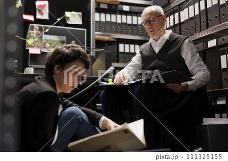 Overworked police officers analyzing criminology report, working late at night at federal criminal investigation in arhive room. Private detectives checking federal files, discussing crime evidence Overworked police officers analyzing criminology report, working late at night at federal criminal investigation in arhive room. Private detectives checking federal files, discussing crime evidence 111325155