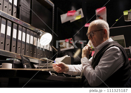 Police officer having remote discussion with inspector, discussing crime scene report using landline phone. Private detective working late at night at criminal case, analyzing crime evidence 111325187