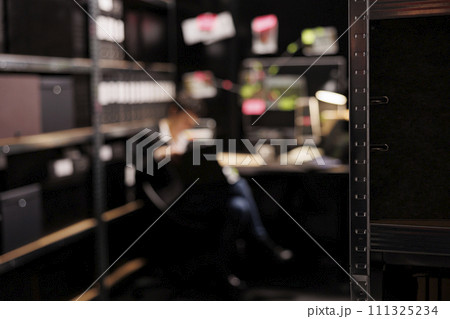 Selective focus of shelves full with evidence files, in background private investigator reading crime scene report. Police officer working late at night at missing person case in arhive room. 111325234