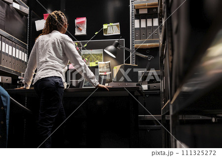 African american woman private detective looking at surveillance photos on laptop and analyzing clues. Investigator standing near desk, studying suspect photographs and evidence 111325272