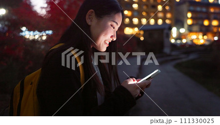 Asian woman, phone and night at city for social media, communication or outdoor networking. Female person relax on mobile smartphone in the late evening for online chatting in urban town of Japan 111330025