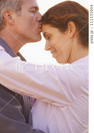Couple, forehead kiss and hug with love, respect or happiness on holiday in retirement. Mature, man and woman embrace together outdoor at sunset on vacation or show affection or gratitude in marriage 111333004