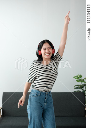 Attractive and happy young Asian woman in casual clothes wearing headphones, relaxes listening to music in her home office. 111334516