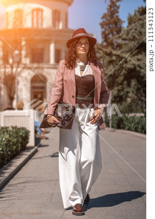 Woman park city. Stylish woman in a hat walks in a park in the city. Dressed in white corset trousers and a pink jacket with a bag in her hands. 111343123