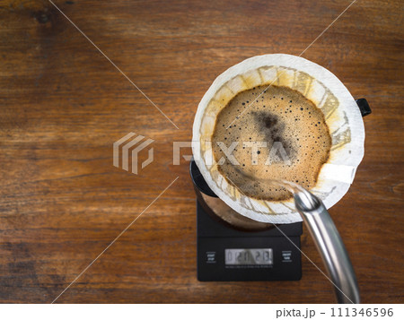 Pouring water on the light roast coffee grounds through the dripper with filter paper. On digital scale, placed on the wooden table. In top view. Pouring water on the light roast coffee grounds through the dripper with filter paper. On digital scale, placed on the wooden table. In top view. 111346596