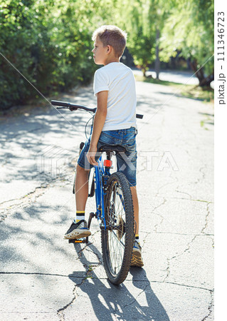 Thoughtful boy on blue bicycle in serene park, enjoying warm weather amidst nature Thoughtful boy on blue bicycle in serene park, enjoying warm weather amidst nature 111346723