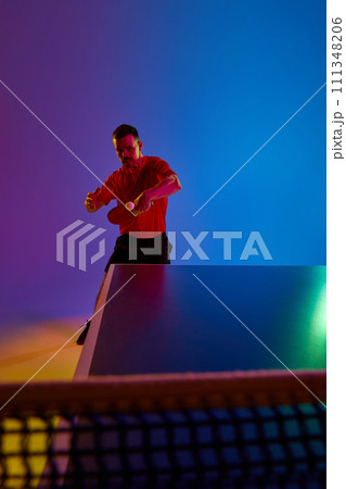 Young man, table tennis competitor in mid-swing during competitive match in neon light against gradient background. Professional sport. Dynamic gel portrait. Young man, table tennis competitor in mid-swing during competitive match in neon light against gradient background. Professional sport. Dynamic gel portrait. 111348206