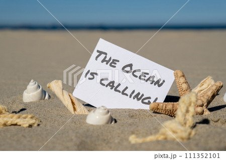 THE OCEAN IS CALLING text on paper greeting card on background of starfish seashell summer vacation decor. Sandy beach sun coast. Holiday concept postcard. Getting away Travel THE OCEAN IS CALLING text on paper greeting card on background of starfish seashell summer vacation decor. Sandy beach sun coast. Holiday concept postcard. Getting away Travel 111352101